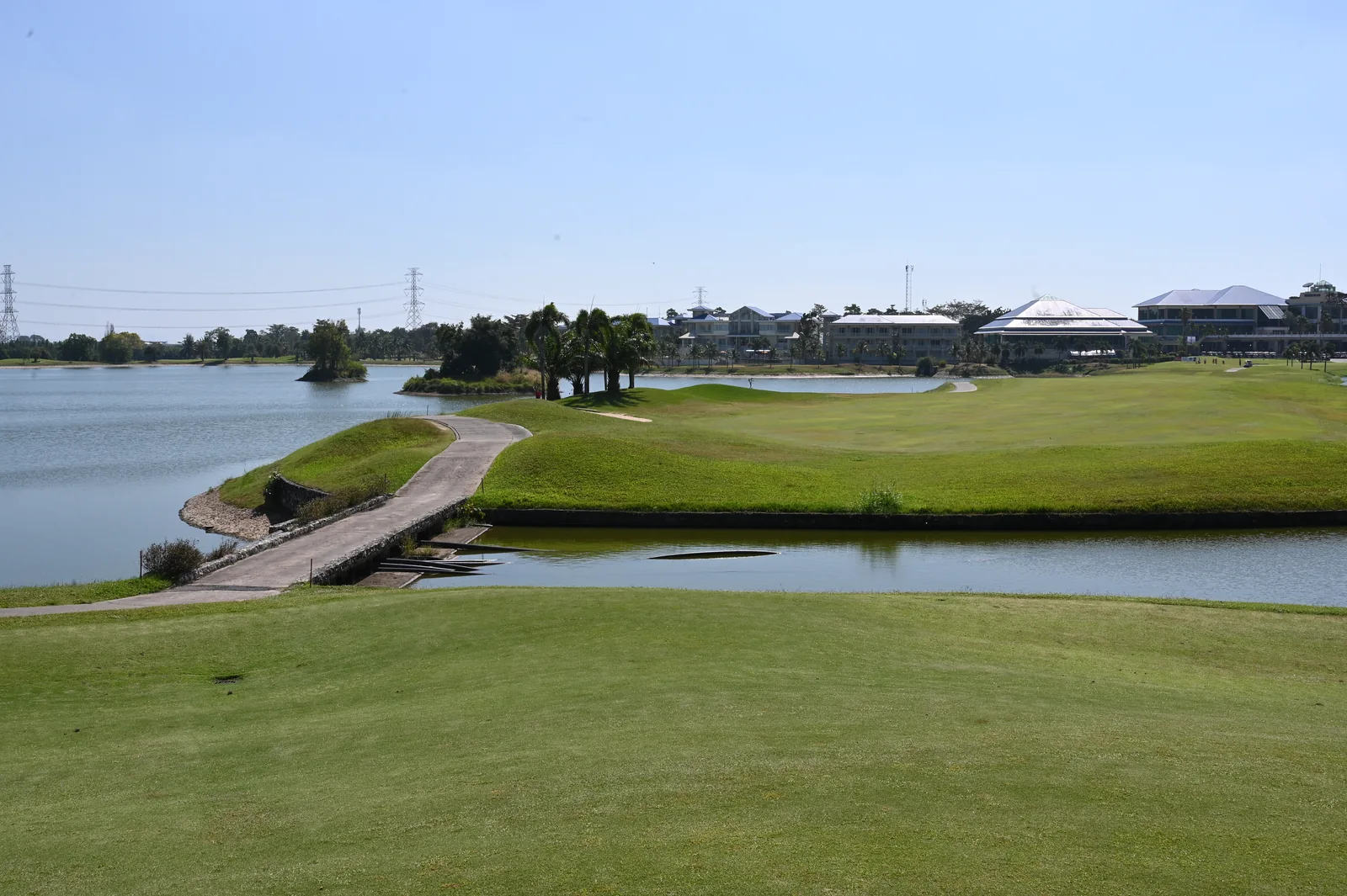 A curved cart path crosses water beside the fairways, with Pattana clubhouse buildings beyond, in December 2024.
