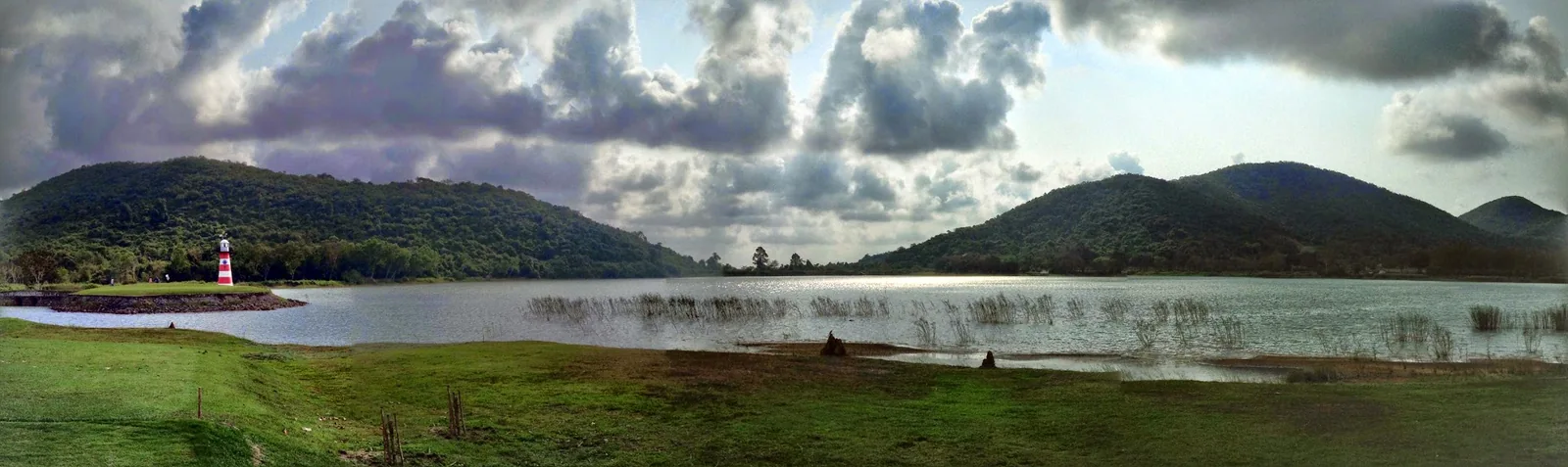 A red-and-white lighthouse beside the Plutaluang golf green, with a broad lake and wooded hills beneath clouds in March 2015.